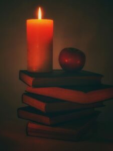 A lit candle and a red apple sitting on a stack of hardcover books in a dimly lit room. 